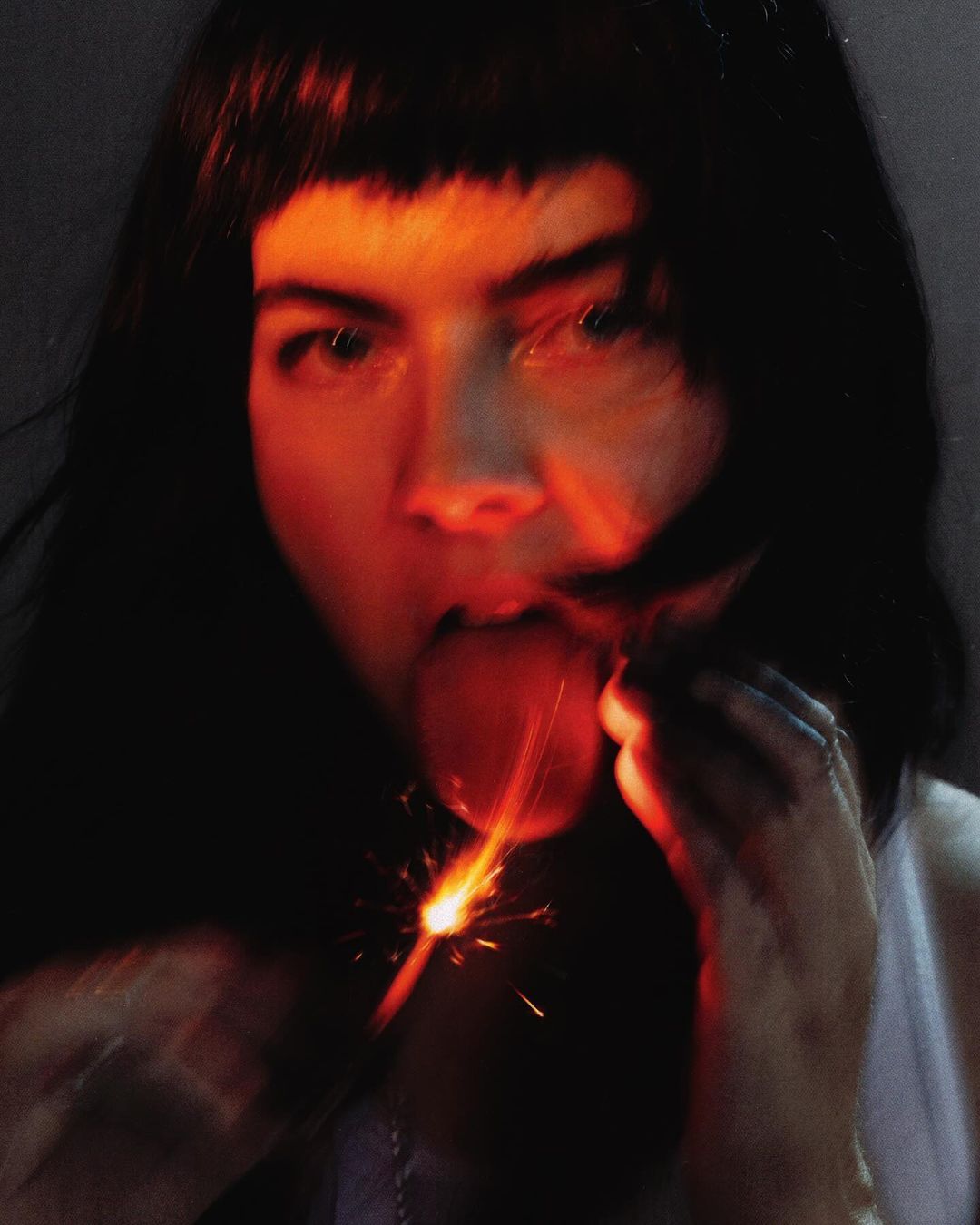 Close-up portrait of a woman with dark hair holding a sparkler near her open mouth, with the background in dark hues.