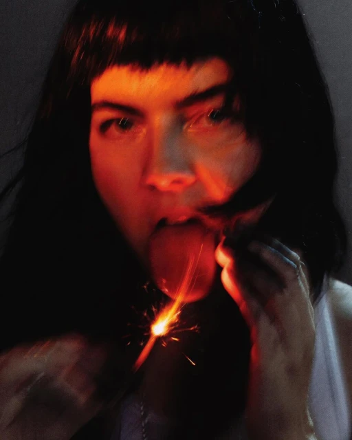 Close-up portrait of a woman with dark hair holding a sparkler near her open mouth, with the background in dark hues.