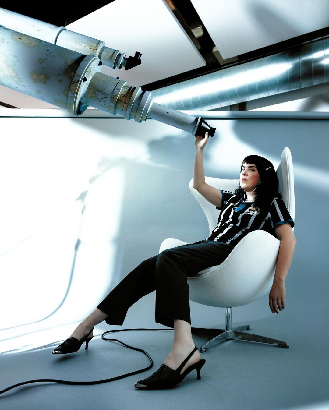 Stylish woman in chair looking through industrial telescope in photo studio. Retro shirt, dark pants, heels, modern white chair, gray floor.