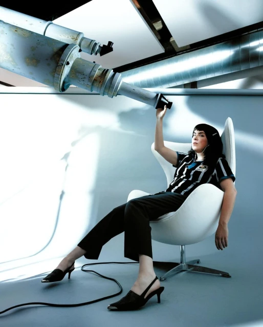 Stylish woman in chair looking through industrial telescope in photo studio. Retro shirt, dark pants, heels, modern white chair, gray floor.