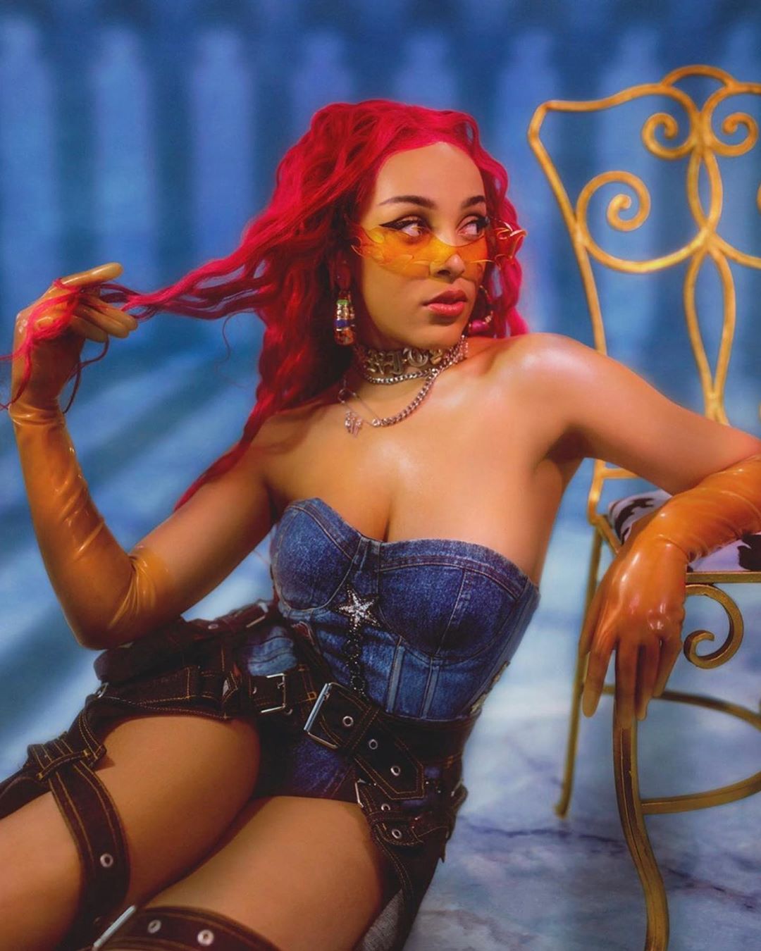 Portrait of woman with red hair, wearing yellow sunglasses and a denim corset, posing in a studio with a golden chair.