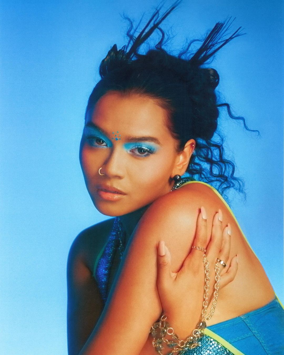 Portrait of a stylish East Asian woman with blue eyeshadow and creative updo, wearing a shimmering blue top against a light blue backdrop.