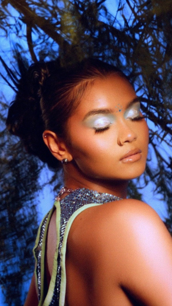 Close-up portrait of a woman with creative silver eyeshadow, dark hair in an updo, and a beaded top against a blue background.
