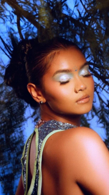 Close-up portrait of a woman with creative silver eyeshadow, dark hair in an updo, and a beaded top against a blue background.