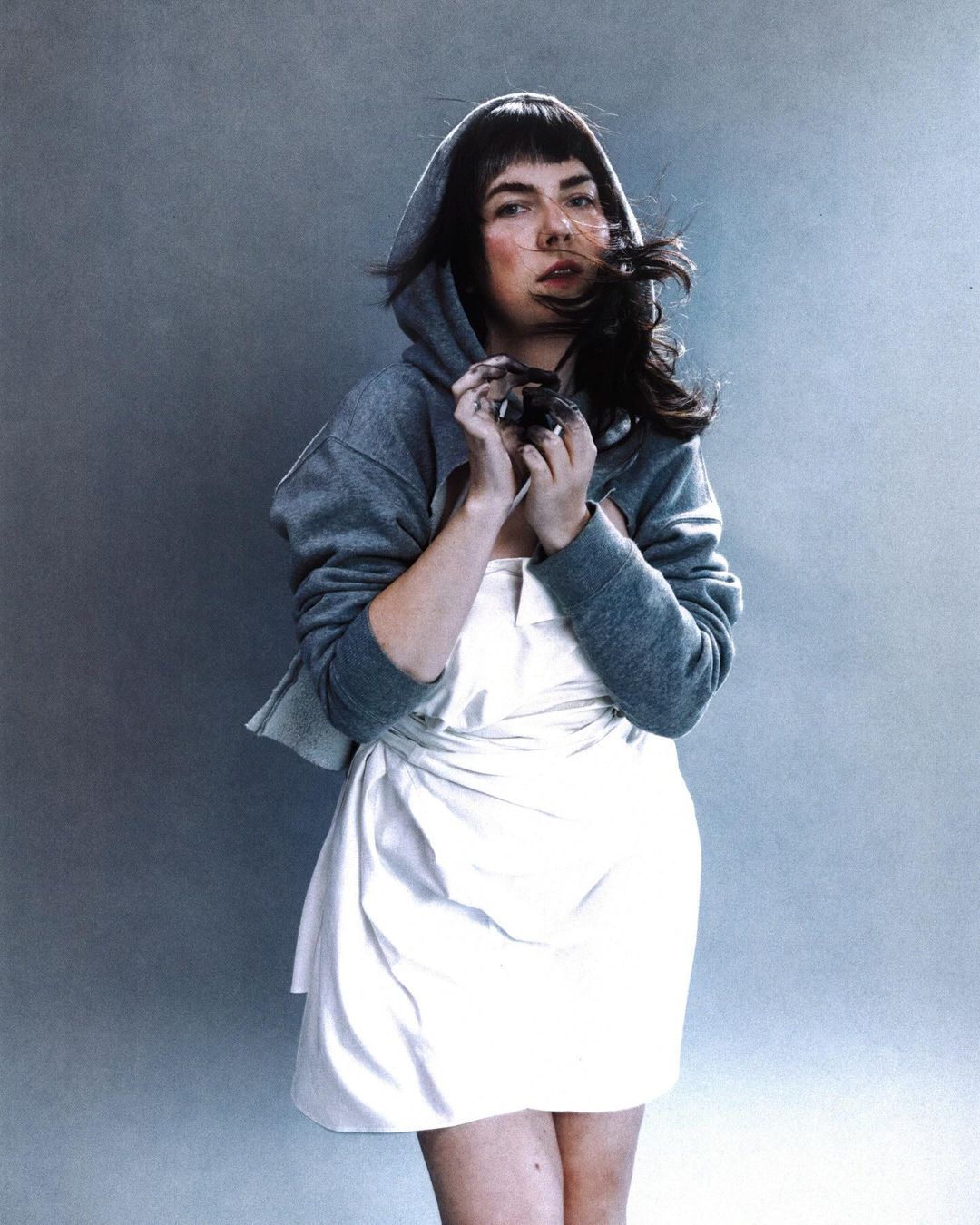 Woman in a hoodie and white dress with hair blowing in the wind, posing in front of a neutral grey background in a studio setting.