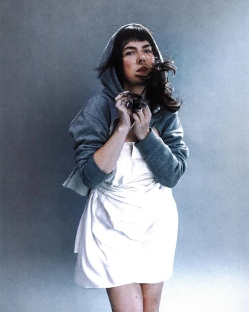 Woman in a hoodie and white dress with hair blowing in the wind, posing in front of a neutral grey background in a studio setting.