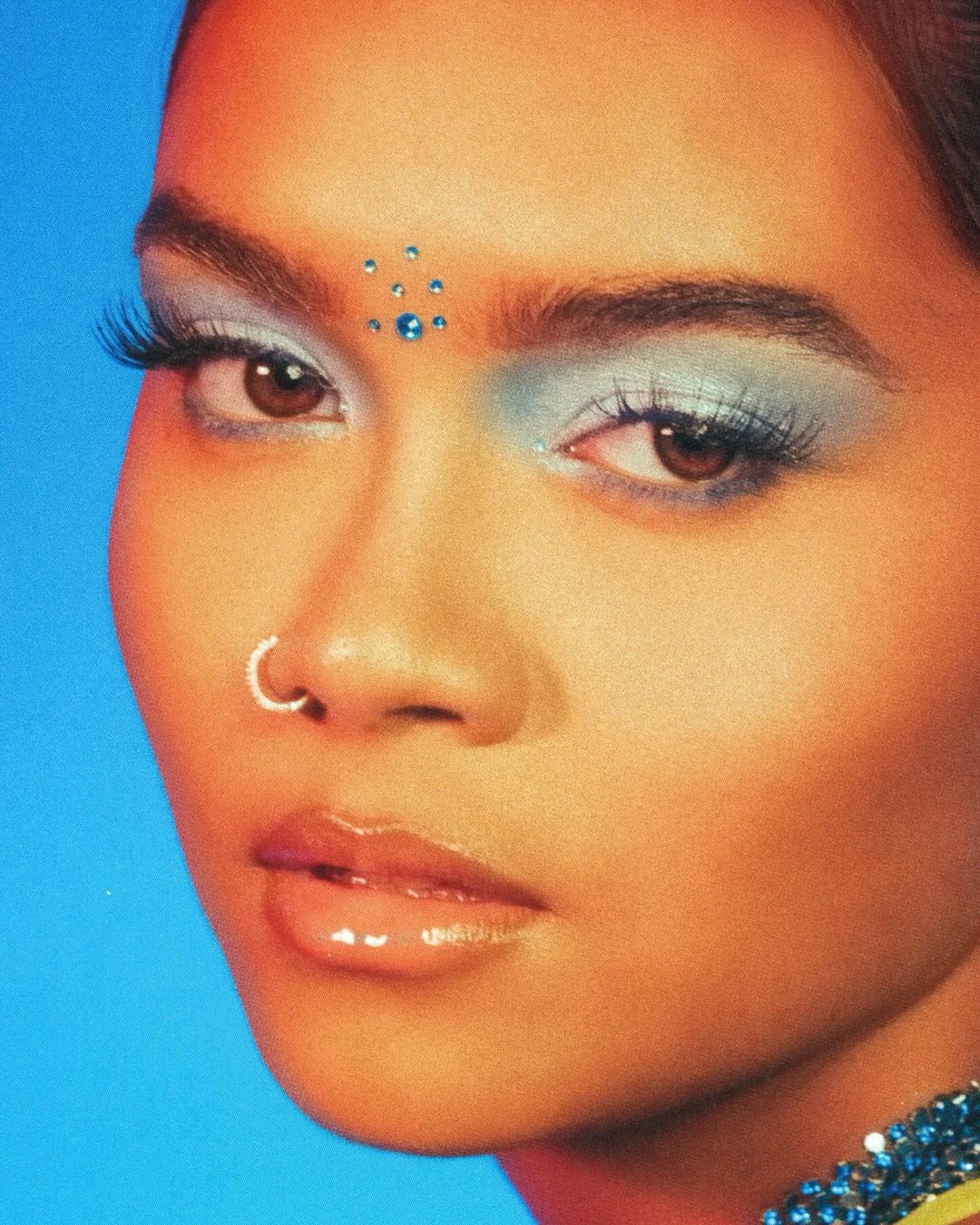 Close-up portrait of a woman with tan skin, light blue eyeshadow, a nose ring, and blue gems on her forehead against a light blue background.