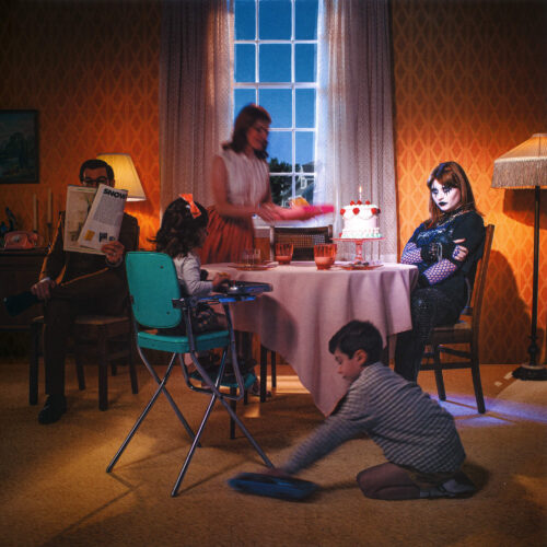 Family gathers around a table for a birthday celebration in an interior setting with a cake, decorations, and dim, moody lighting.