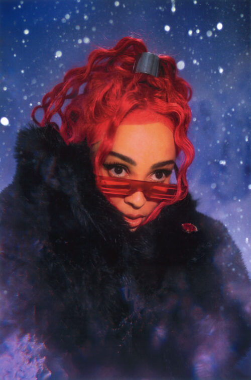 Close-up of a person with red curly hair, wearing red tinted sunglasses and a black furry coat against a blue, starry background.