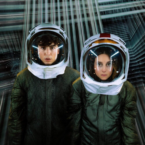 Two young people in space suits with helmets against a futuristic, patterned backdrop, looking directly at the camera with serious expressions.