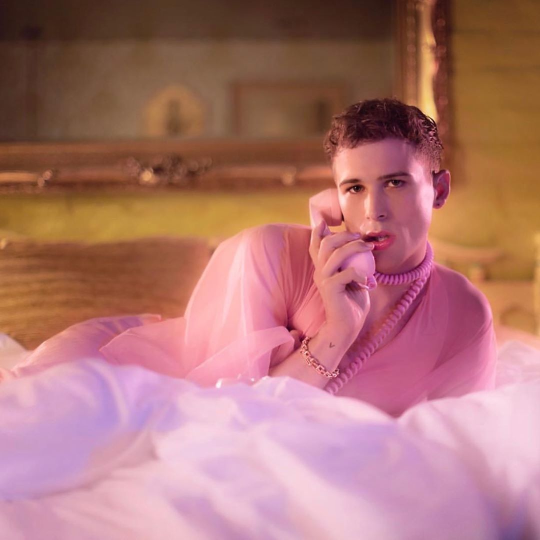 Adult male with short brown hair, wearing pink robe and necklace, lies on a bed using a pink retro-style telephone in an indoor setting.