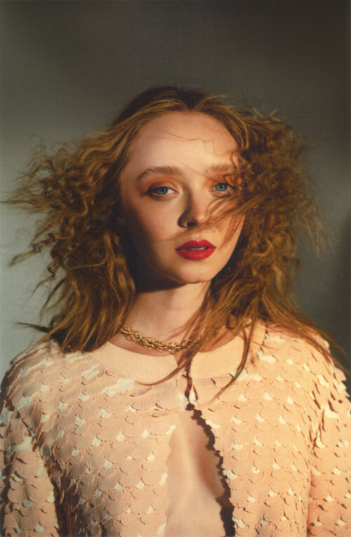 Close-up portrait of a curly-haired adult female wearing a peach sequined jacket and gold necklace, set against a neutral background.