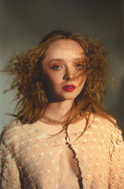 Close-up portrait of a curly-haired adult female wearing a peach sequined jacket and gold necklace, set against a neutral background.