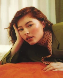 Close-up portrait of a woman with red hair wearing an olive green jacket and polka dot scarf, reclining on a red surface.