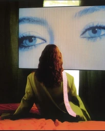 Person with long brown hair wearing an olive green jacket sitting in front of a glowing screen displaying giant blue eyes