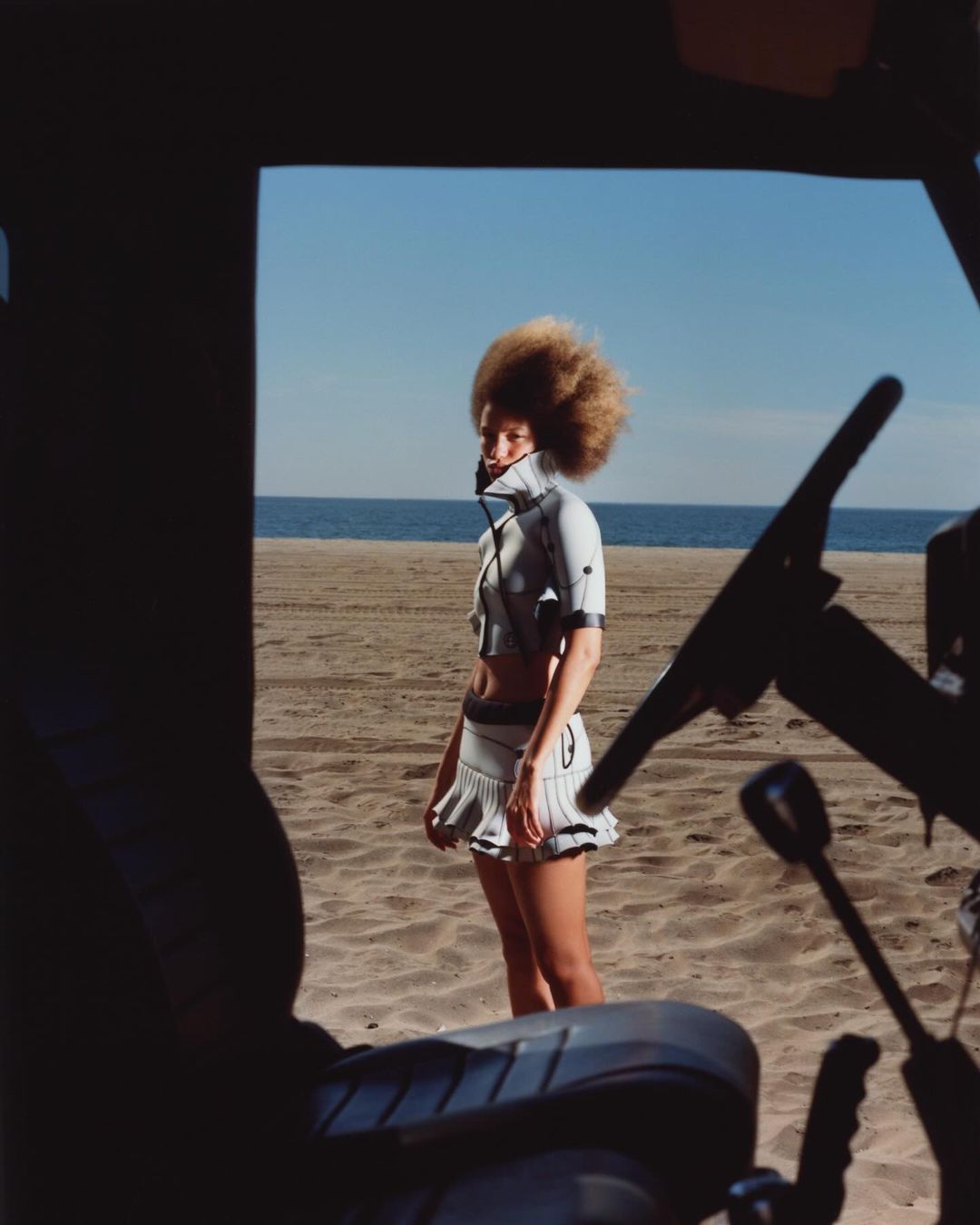 A woman with an afro stands on a sandy beach in a futuristic outfit with the ocean and sky in the background, framed by vehicle elements.