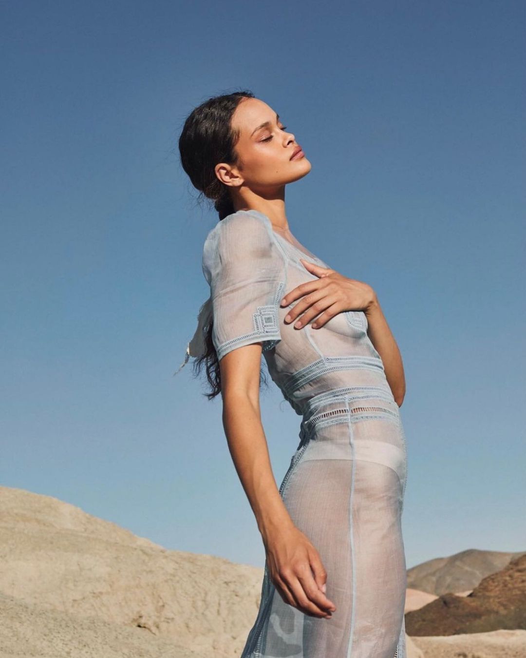 Young woman poses outdoors in sheer blue dress against sky and desert backdrop, showcasing fashion and natural light portraiture.