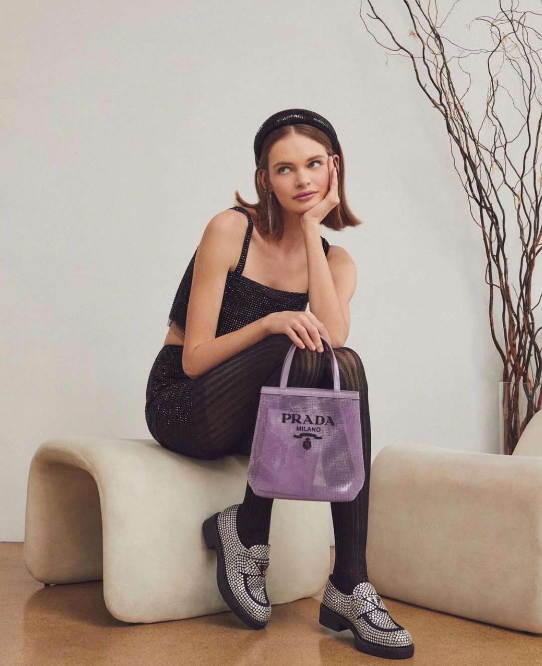 Woman in sparkly top and leggings sits holding a purple Prada tote bag, paired with embellished loafers, against a neutral backdrop.