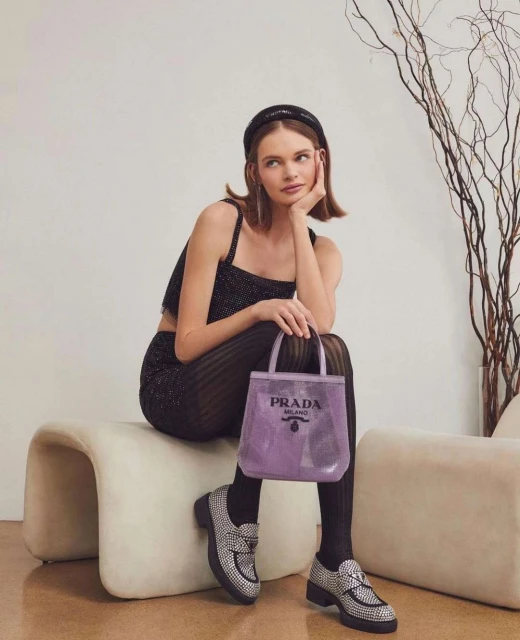 Woman in sparkly top and leggings sits holding a purple Prada tote bag, paired with embellished loafers, against a neutral backdrop.