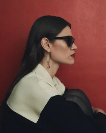 Profile portrait of a woman wearing dark sunglasses, oversized hoop earrings, and a two-tone top against a red background.