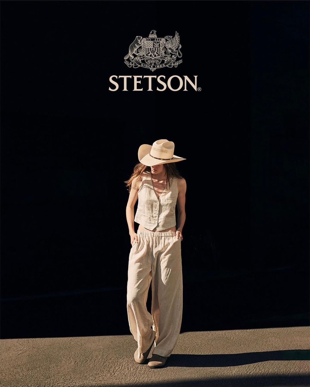 Woman in a linen vest and pants, wearing a cowboy hat, poses in front of a black wall with a Stetson logo, bathed in natural light.