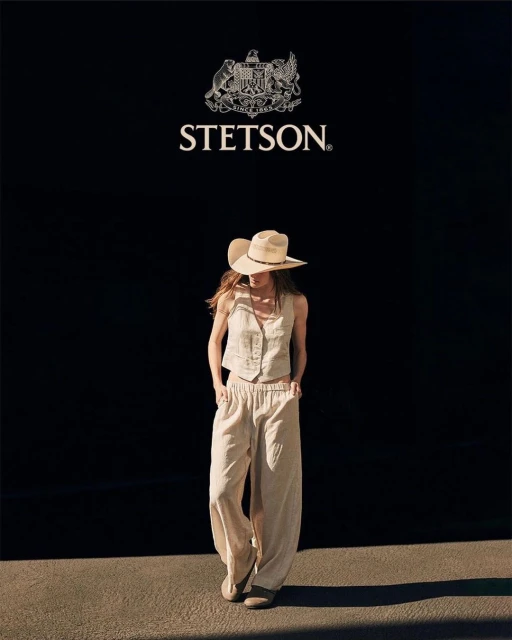 Woman in a linen vest and pants, wearing a cowboy hat, poses in front of a black wall with a Stetson logo, bathed in natural light.