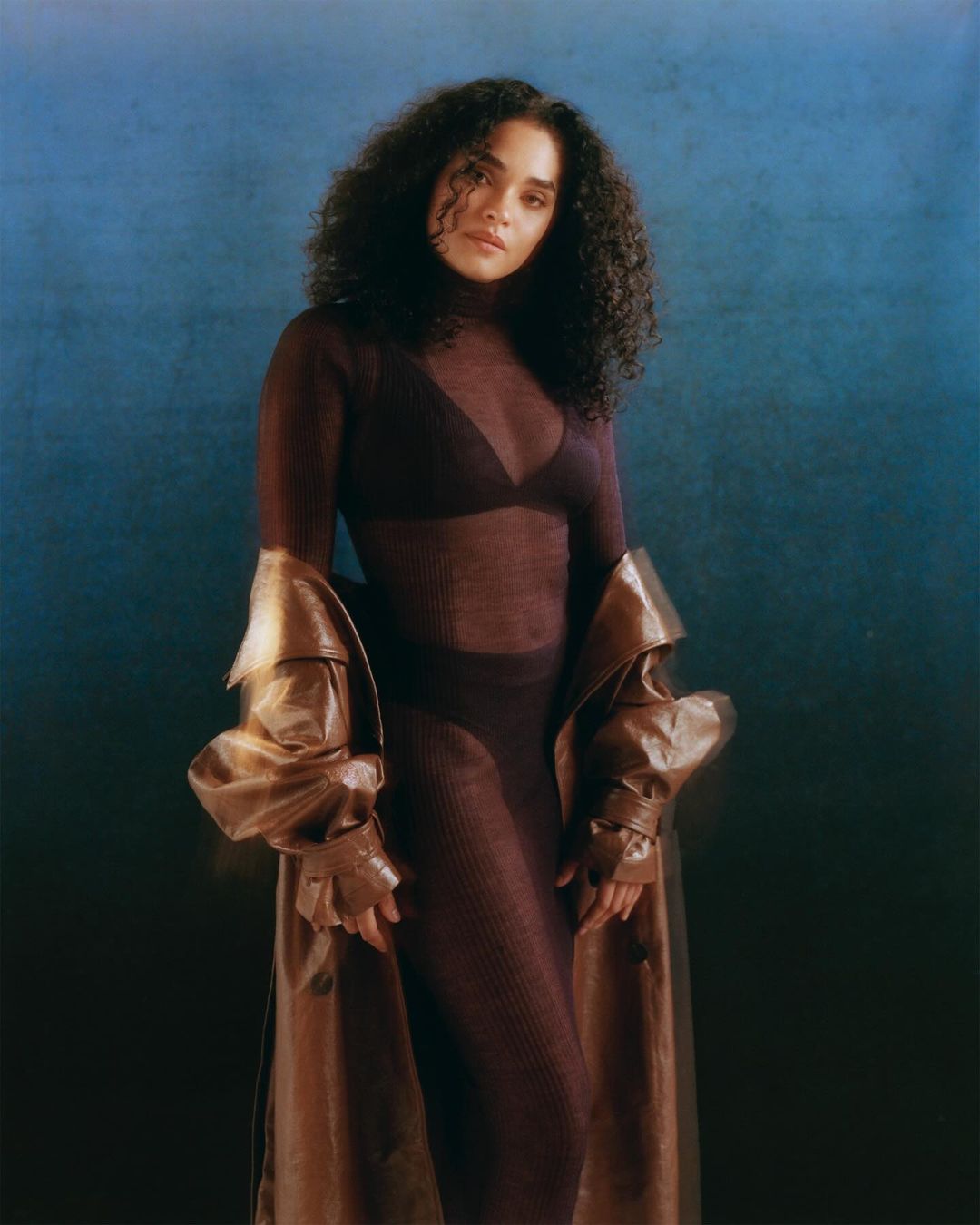 Elegant studio portrait of a mixed-race woman with curly hair, wearing a sheer top, pants, and a brown trench coat against a blue backdrop.