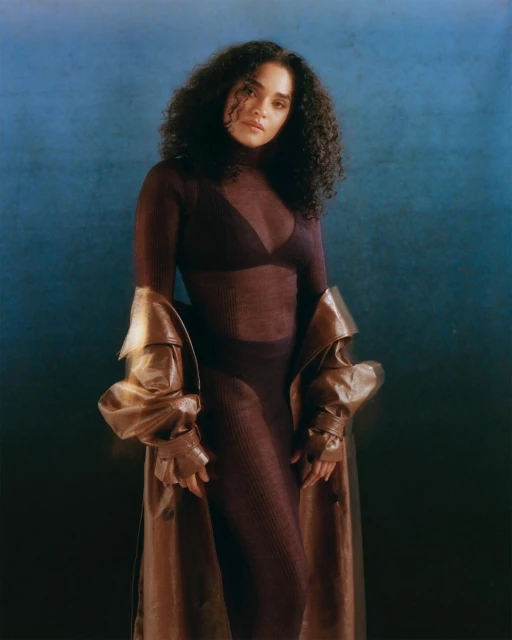 Elegant studio portrait of a mixed-race woman with curly hair, wearing a sheer top, pants, and a brown trench coat against a blue backdrop.