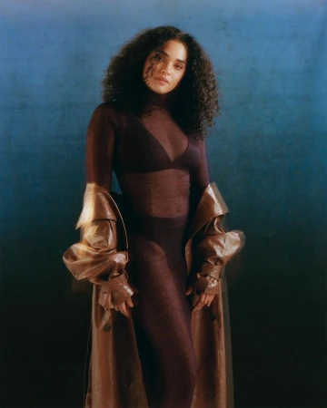 Elegant studio portrait of a mixed-race woman with curly hair, wearing a sheer top, pants, and a brown trench coat against a blue backdrop.