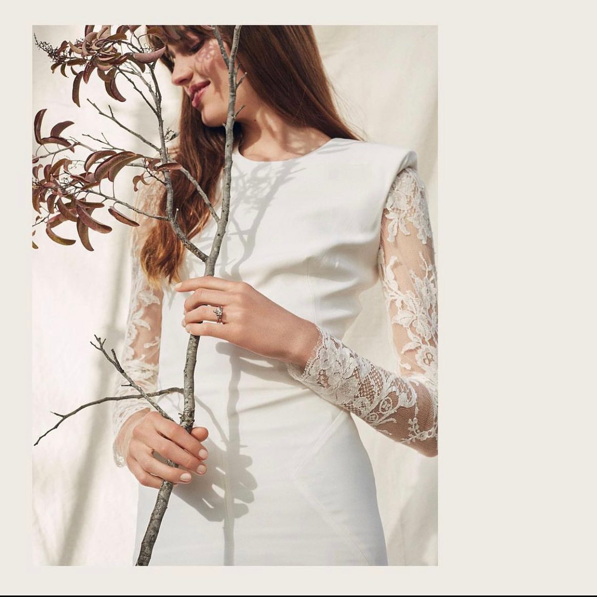Woman in a white long-sleeve dress posing with a branch in front of an off-white backdrop, soft lighting, fashion shoot.