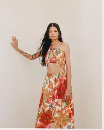 Woman with long dark hair wearing a two-piece floral outfit posing against a plain off-white wall.