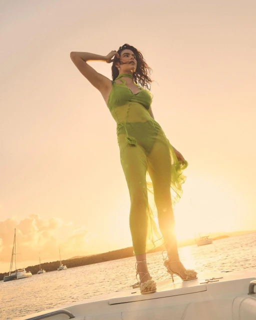 A model poses on a boat at sunset, wearing a green dress and strappy heels, with the sun and other boats visible in the background.