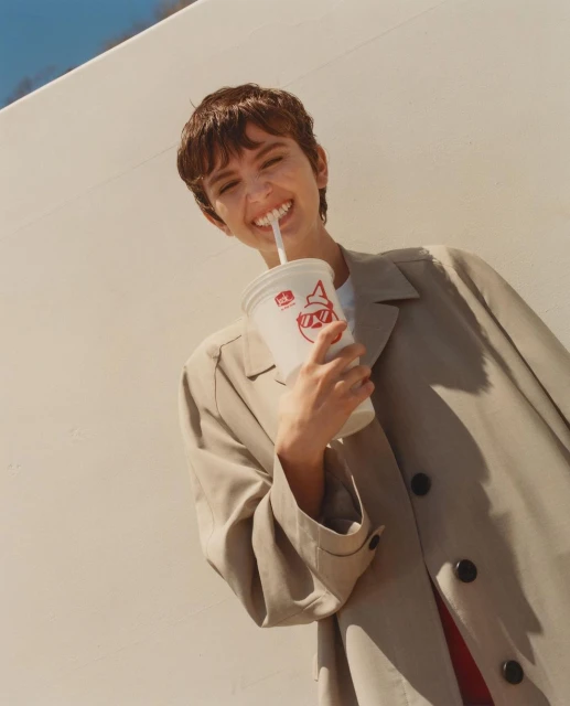 Young woman in a tan trench coat smiles while drinking a beverage with a straw outdoors against a light-colored wall in sunlight.
