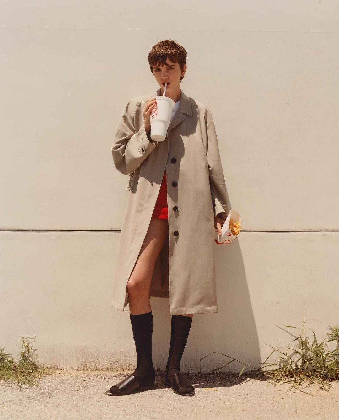 Woman in trench coat sips from a drink while holding fries. She stands against a wall in fashion shot with natural outdoor lighting.