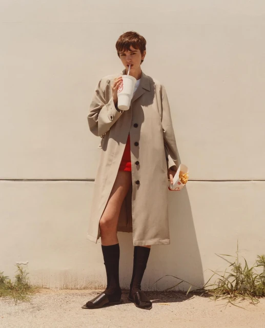 Woman in trench coat sips from a drink while holding fries. She stands against a wall in fashion shot with natural outdoor lighting.