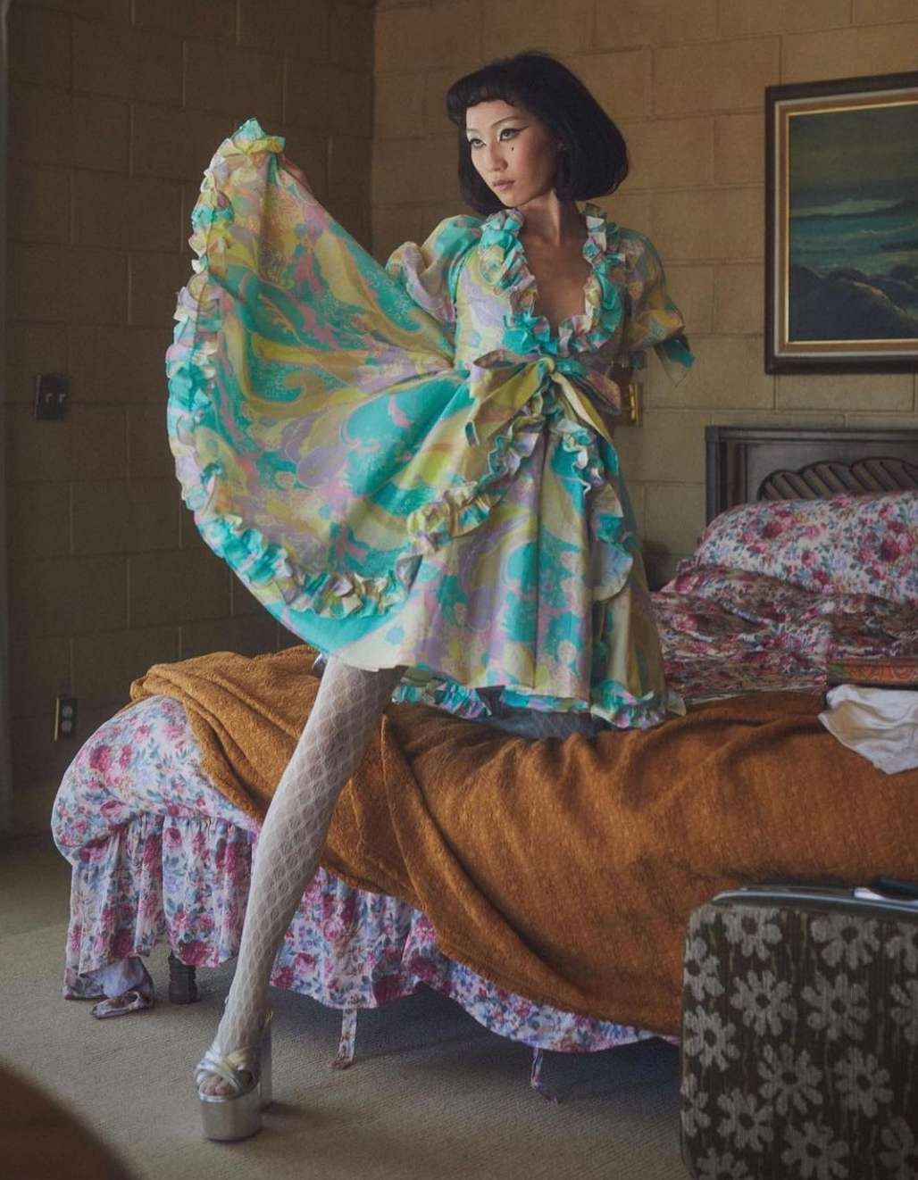 Woman poses indoors wearing a multicolor ruffle dress, white patterned tights, and platform shoes. She is standing near a bed.