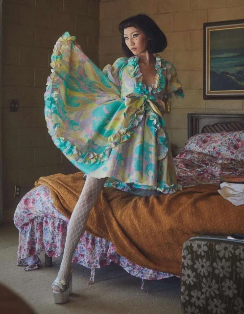 Woman poses indoors wearing a multicolor ruffle dress, white patterned tights, and platform shoes. She is standing near a bed.