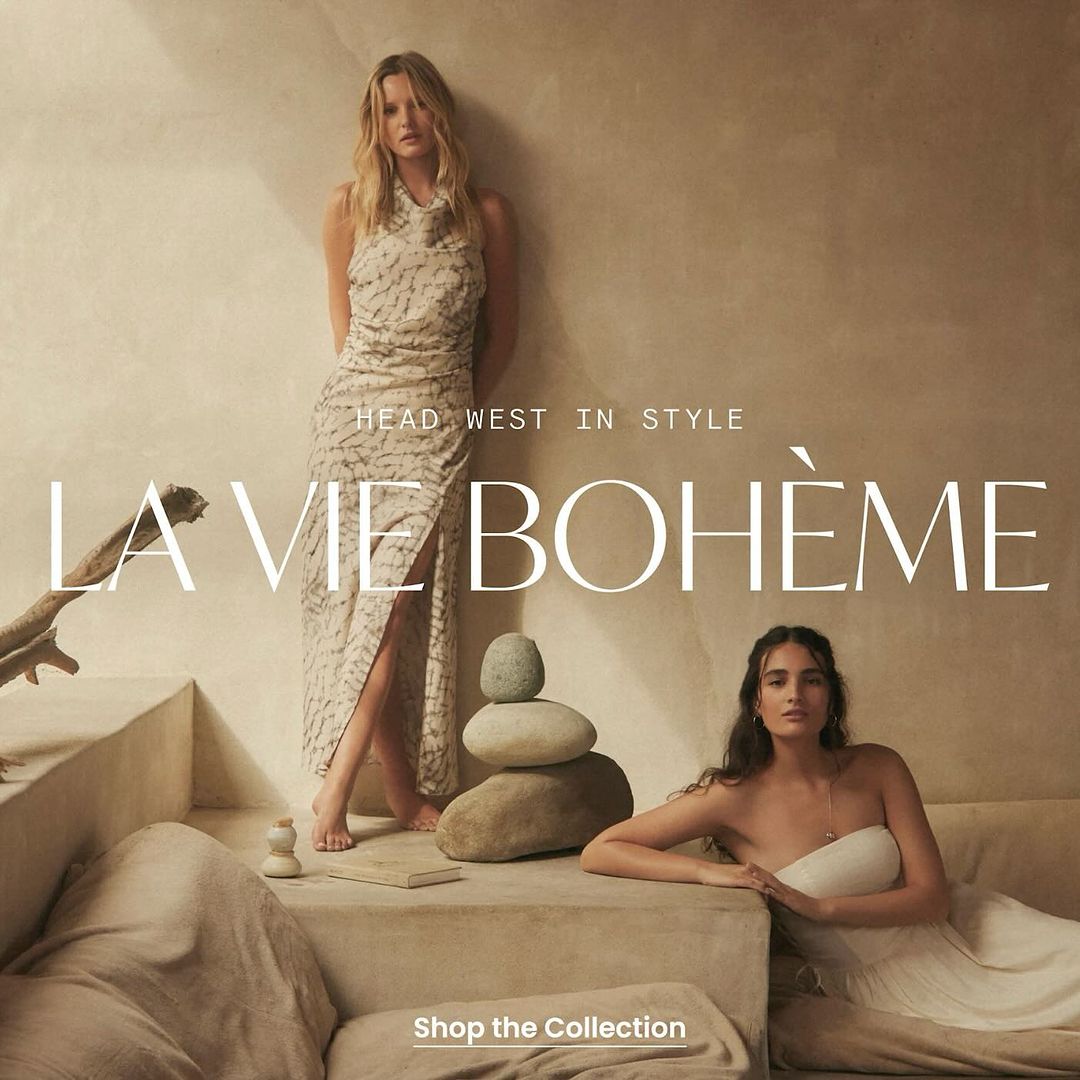 Two women pose in neutral-toned dresses. One woman stands next to stacked stones, and another reclines on a draped stone sofa indoors.
