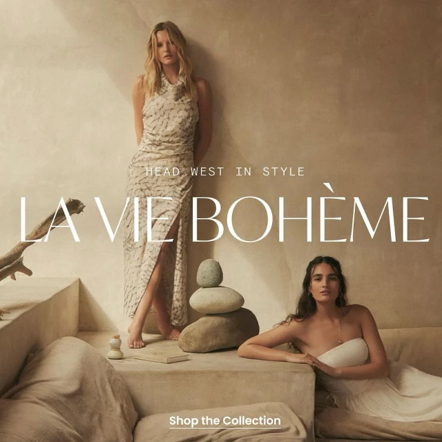 Two women pose in neutral-toned dresses. One woman stands next to stacked stones, and another reclines on a draped stone sofa indoors.