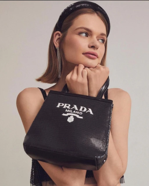 Woman posing with a black Prada sequin tote bag, wearing a matching headband and dangling earrings against a neutral backdrop.