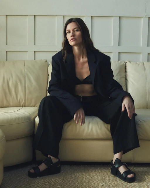 Brunette woman in a dark bra, oversized blazer, and wide-leg trousers sits on a beige couch in an indoor setting, looking at the camera.