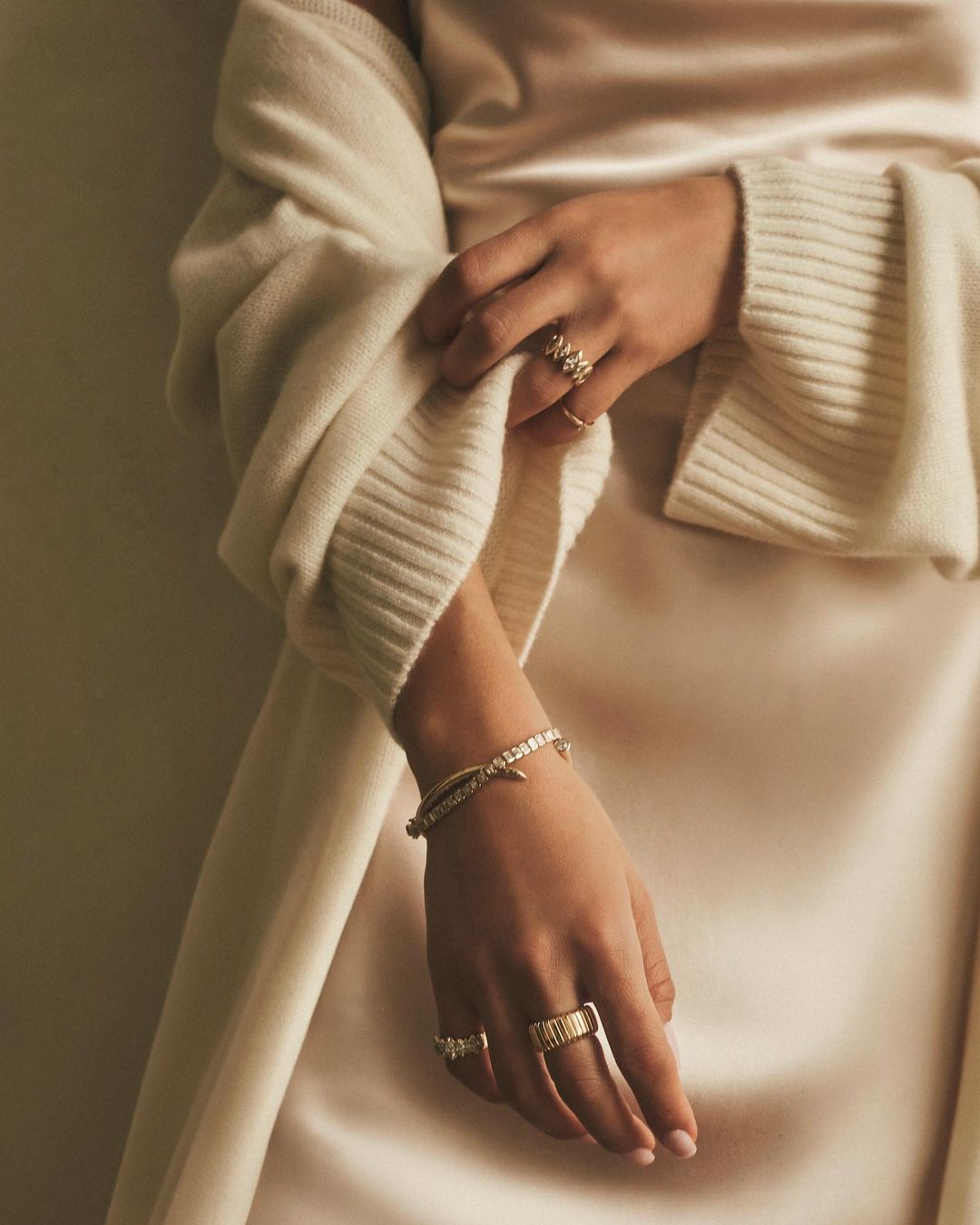 Close-up of a woman's hands adorned with gold rings and a bracelet, elegantly posed against a backdrop of a cream-colored dress and sweater.