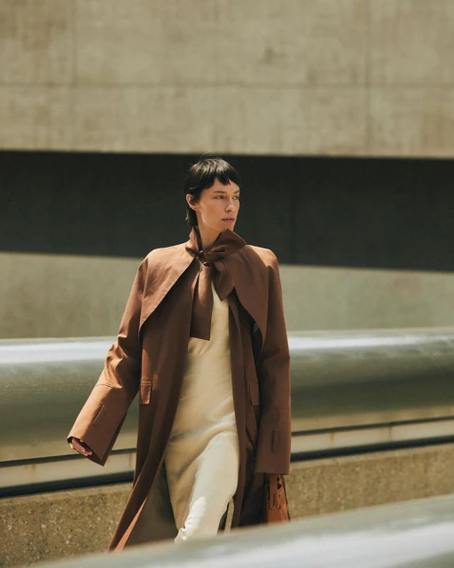 Woman in a brown trench coat and white dress walking in an urban setting with concrete walls and metal railing, fashion editorial.