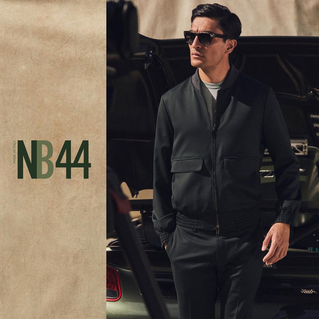 Man wearing dark sunglasses and a black bomber jacket standing in front of a luxury sports car, photographed by Justin Wilczynski.