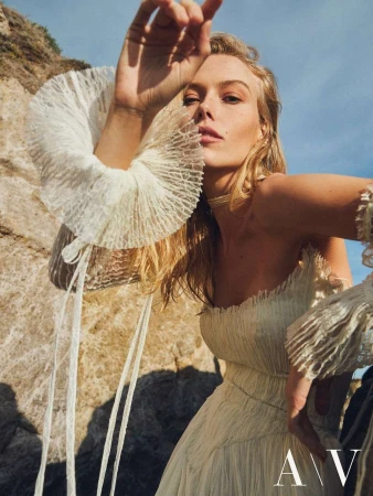 Blonde woman in an off-shoulder white textured dress with ruffled cuffs posing outdoors against rocky coastal cliffs under a bright blue sky.