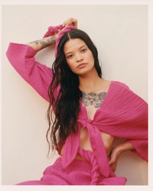 Portrait of an adult female in a pink textured outfit holding a matching pink rope headband, posing in front of a neutral-toned wall.