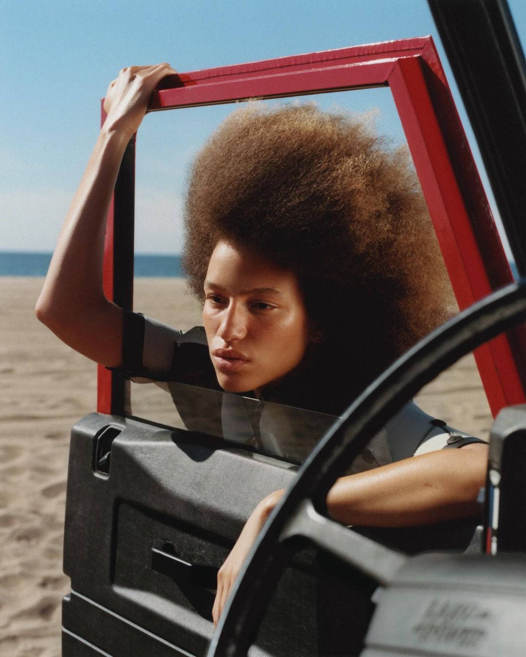 A woman with an afro poses by the open door of a car on a beach, framed by the red door frame. The sky is blue and the sand is neutral.