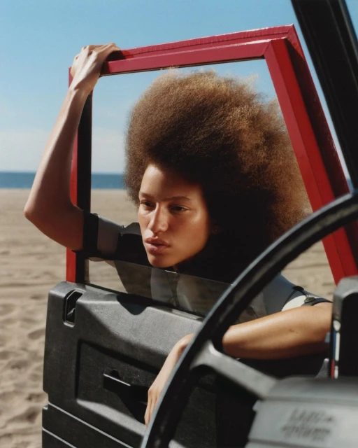 A woman with an afro poses by the open door of a car on a beach, framed by the red door frame. The sky is blue and the sand is neutral.