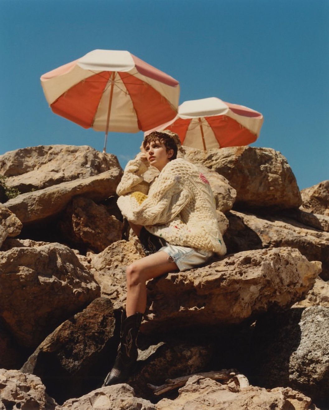 Young adult woman in a sweater and boots poses on rocks under two parasols against a blue sky on a sunny day in an outdoor setting.