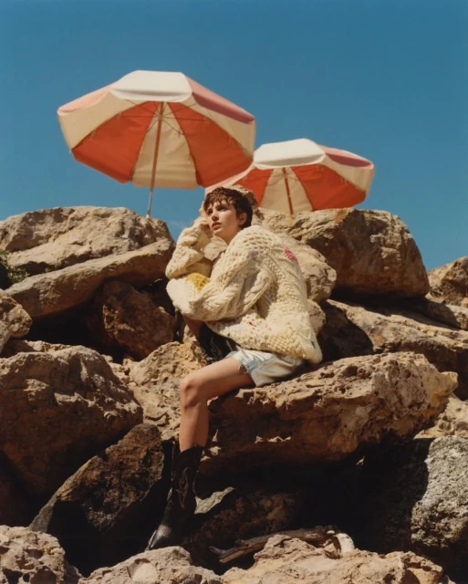 Young adult woman in a sweater and boots poses on rocks under two parasols against a blue sky on a sunny day in an outdoor setting.